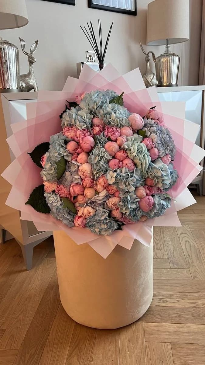 Pale blue hydrangeas and pink peonies wrapped in layered pink paper, standing on a velvet pouf