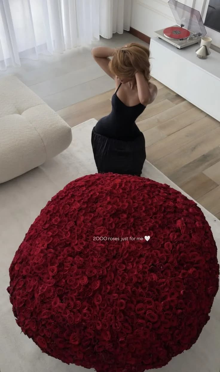 A huge dome of deep red roses on a living room rug, with a woman in a black dress kneeling behind it