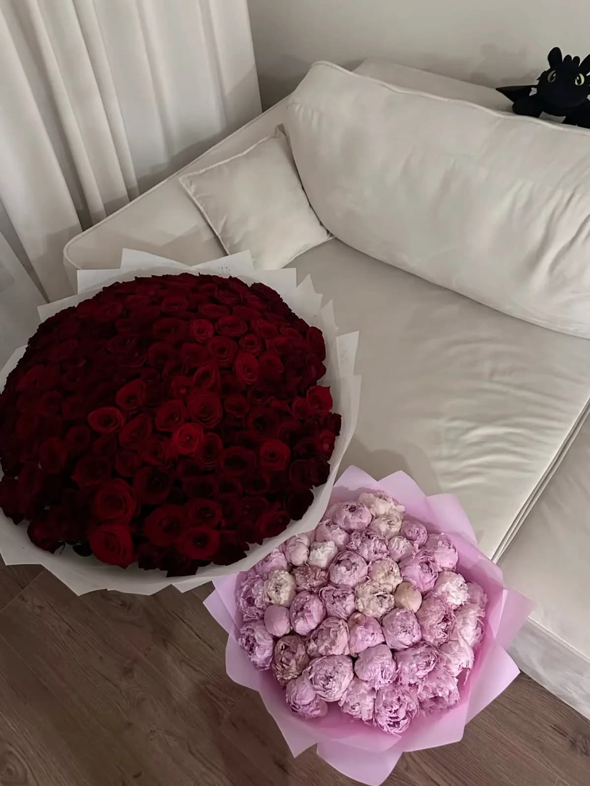 A large bouquet of red roses in white wrap beside a bouquet of pink peonies in pink wrap, set on a cream sofa